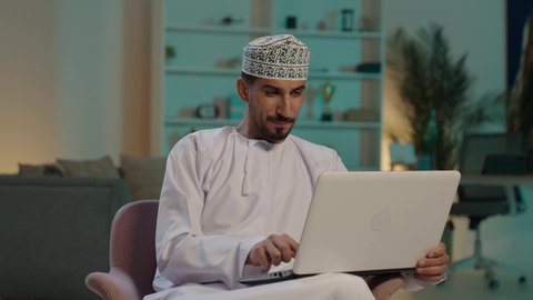 التواصل مع الأهل والأصدقاء، دمج العمل مع التقنية والتكنولوجيا، مهن ووظائف ادارية مكتبية, إدارة شؤون الأعمال وتنظيمها عبر الاجهزة التقنية، ايماءات السعادة والسرور بتحقيق الأهداف، تصوير  لرجل عربي خليجي عماني يرتدي الدشداشة والكمة  يحمل بيده جهاز الحاسوب المحمول