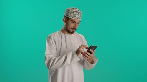 تكبير الصورة لمشاهدة التفاصيل، مشاهدة الأفلام والمسلسلات الممتعة، متابعة مواقع التواصل الاجتماعي عبر الهاتف المحمول، التواصل مع الأهل والأصدقاء، استخدام التكنولوجيا و التقنيات الحديثة, تصوير لرجل عربي خليجي عماني يرتدي الدشداشة والكمه يحمل بيده جهاز الجوال، خلفية زرقاء