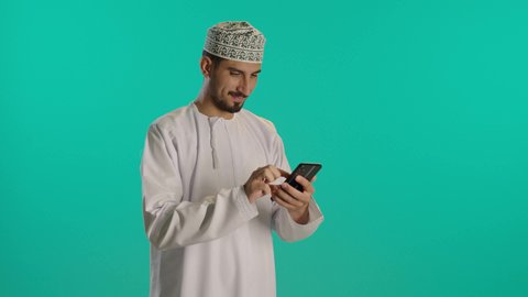 متابعة مواقع التواصل الاجتماعي عبر الهاتف المحمول، مشاهدة الأفلام والمسلسلات الممتعة، التواصل مع الأهل والأصدقاء، استخدام التكنولوجيا و التقنيات الحديثة, تصوير لرجل عربي خليجي عماني يرتدي الدشداشة والكمه يحمل بيده جهاز الجوال، خلفية زرقاء