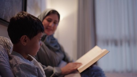الأنشطة التعليمية وتنميه قدرات الابناء بالتعلم، اهتمام الأم بالابناء، قراءة القصص والروايات، ام تقضي وقت ممتع مع الابن قبل النوم، امرأة عربية خليجية سعودية ترتدي العباءة و الحجاب تقرأ لإبنها قصه مسلية، مفهوم السعادة الاسرية