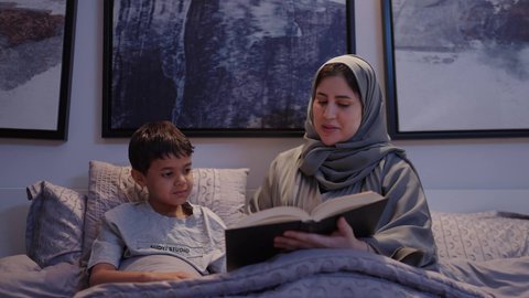 The mother's interest in her children, reading stories and novels, a mother spending enjoyable time with her son before bedtime, an Arab Gulf Saudi woman wearing an abaya and hijab reading an entertaining story to her son, the concept of family happiness, educational activities, and developing children's abilities through learning.