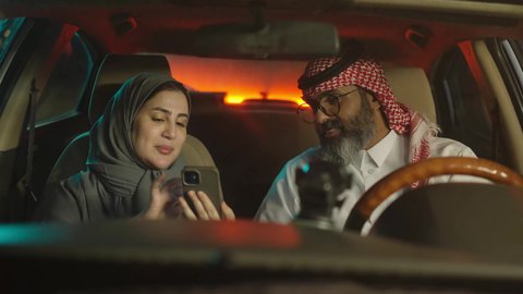 Spending enjoyable family time, a Saudi Arabian couple sitting inside the car using their mobile phones, discussing and exchanging opinions, having a good time outdoors.
