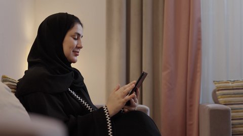 النظر للكاميرا بايماءات السعادة و السرور، قضاء وقت ممتع في المنزل، استخدام التقنية الحديثة، الدردشة مع الاهل والاصداقاء، امراة عربية خليجية سعودية تجلس فى غرفه المعيشة تقوم بتصفح الانترنت عبر الهاتف المحمول