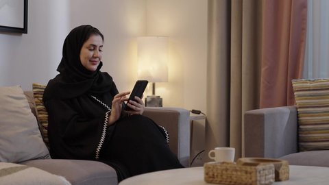 الدردشة مع الاهل والاصداقاء، استخدام التقنية الحديثة، امراة عربية خليجية سعودية تجلس فى غرفه المعيشة تقوم بتصفح الانترنت عبر الهاتف المحمول, النظر للكاميرا بايماءات السعادة و السرور، قضاء أوقات ممتعة في المنزل