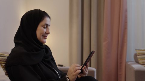 قضاء وقت ممتع في المنزل، استخدام التقنية الحديثة، الدردشة مع الاهل والاصداقاء، امراة عربية خليجية سعودية تجلس فى غرفه المعيشة تقوم بتصفح الانترنت عبر الهاتف المحمول
