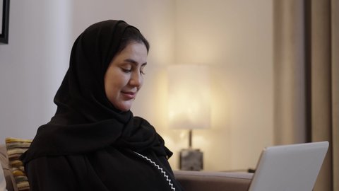 دمج العمل مع التقنية والتكنولوجيا، إدخال البيانات والكتابة من خلال لوحة المفاتيح، امتلاك مهارة الكتابة السريعة على الكيبورد، تصوير من الجانب لإمرأة عربية خليجية سعودية محجبة ترتدي العباءة تجلس فى غرفه المعيشة تقوم بالعمل على الحاسب المحمول