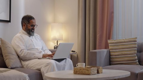 Communicating with family and friends via laptop, completing remote work tasks with integration gestures, managing and organizing business affairs, using a modern and advanced technological device, an Arab Gulf Saudi man wearing a white thobe using a laptop.