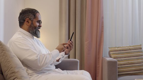 Communication and chatting with friends and relatives via mobile, spending enjoyable time at home, using modern technological devices, expressions and gestures of joy and happiness, a side shot of a Gulf Arab Saudi man wearing a white thobe holding a mobile phone sitting in the living room.