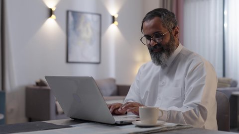إنجاز مهام العمل عن بعد، استخدام جهاز تقني حديث ومتطور، إدارة شؤون الأعمال وتنظيمها عبر اللابتوب، الاستمتاع بالمشروبات الساخنة في المنزل، رجل عربي خليجي سعودي يرتدي الثوب الابيض يجلس فى غرفة المعيشة يستخدم الحاسوب المحمول