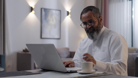 Using a modern and advanced technical device, managing and organizing business affairs via a laptop, enjoying hot beverages at home, an Arab Gulf Saudi man wearing a white thobe sitting in the living room using a laptop, completing work tasks remotely.