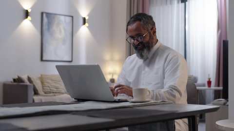 Managing work affairs and organizing them via laptop, enjoying a hot drink at home, using a modern and advanced technological device, an Arab Gulf Saudi man wearing a white thobe sitting in the living room using a laptop, completing remote work tasks.