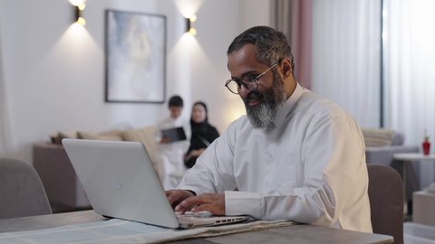 Using a tablet device, the concept of fatherhood and great love, remote work, an Arab Gulf Saudi man wearing a white thobe sitting with his wife and son working on a laptop.