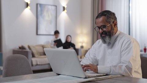 مفهوم الأبوة والحب الكبير، استخدام جهاز التابلت، العمل عن بعد، رجل عربي خليجي سعودي يرتدي الثوب الابيض يجلس برفقة زوجته و ابنه يقوم بالعمل على الحاسب المحمول