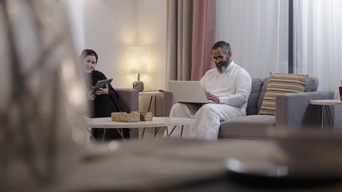 التعلم عن بعد وتلقي الدروس عبر الأونلاين، استخدام جهاز التابلت، رجل عربي خليجي سعودي يرتدي الثوب الابيض يجلس برفقة زوجته يقوم بالعمل على الحاسب المحمول