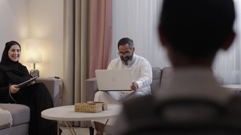 جو عائلي سعيد، تعابير الحب والعطف على الابن، مفهوم الرابطة الأبوية والترابط الأسري، استخدام جهاز التابلت، رجل عربي خليجي سعودي يرتدي الثوب الابيض يجلس برفقة زوجته يقوم بتقبيل ابنه و يمسك بيدية الحاسب المحمول