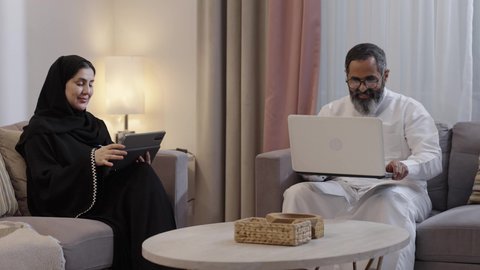 تعابير الحب والعطف على الابن، مفهوم الرابطة الأبوية والترابط الأسري، استخدام جهاز التابلت، رجل عربي خليجي سعودي يرتدي الثوب الابيض يجلس برفقة زوجته يقوم بتقبيل ابنه و يمسك بيدية الحاسب المحمول