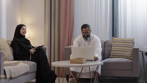 The concept of parental bond and family connection, using a tablet device, an Arab Gulf Saudi man wearing a white thobe sitting with his wife, kissing his son and holding a laptop in his hands.
