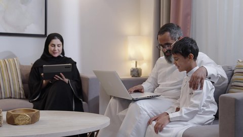 القيام  بنشاط مسلي في المنزل، قضاء وقت عائلى ممتع، الاشارة الى شاشة الحاسوب، الاهتمام و الرعاية بالابن ،استخدام جهاز التابلت، رجل عربي خليجي سعودي يرتدي الثوب الابيض يجلس برفقة زوجته و ابنه يعمل على الحاسب المحمول