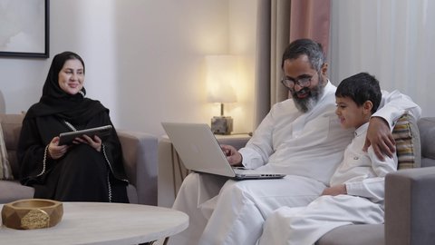 قضاء وقت عائلى ممتع، الاشارة الى شاشة الحاسوب، الاهتمام و الرعاية بالابن ،استخدام جهاز التابلت، رجل عربي خليجي سعودي يرتدي الثوب الابيض يجلس برفقة زوجته و ابنه يعمل على الحاسب المحمول