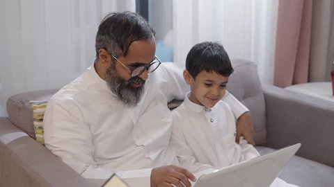 Referring to the computer screen, caring for and nurturing the son, spending free time learning and self-educating, an Arab Gulf Saudi man wearing a white thobe sits with his wife and son working on a laptop, enjoying quality family time.