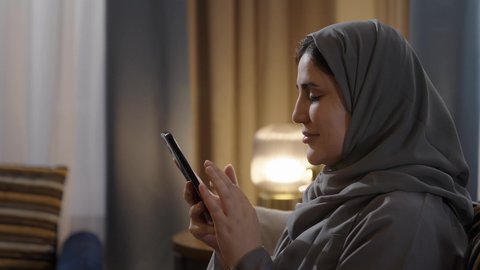 Social interaction through social media, using modern technological devices, a side shot of a Gulf Arab Saudi woman wearing an abaya and hijab sitting in the living room holding a mobile phone, gestures of happiness and joy.