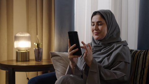 الدردشة مع الأهل والأصدقاء وتصفح مواقع التواصل الاجتماعي، استخدام الأجهزة التقنية الحديثة، تصوير من الجانب لامراة عربيه خليجيه سعودية ترتدي العباءة و الحجاب تجلس في غرفة المعيشة تمسك الهاتف المحمول، ايماءات السعادة و السرور