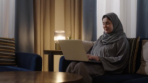 Typing on the keyboard, shopping through websites, working remotely, and browsing the internet using modern technologies, an Arab Gulf Saudi woman wearing a hijab and an abaya sits in the living room using a laptop, chatting, socializing, and making a video call.