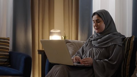 Online shopping, remote work, and browsing the internet using modern technologies. A Saudi Arabian Gulf woman wearing a hijab and an abaya sits in the living room using a laptop, chatting, social networking, making a video call, and typing on the keyboard.