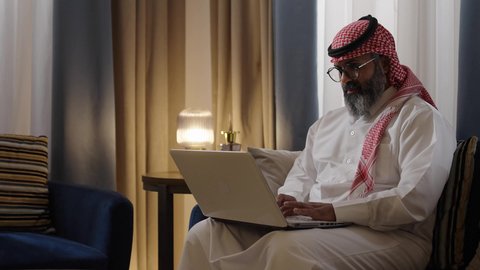 ايماءات السعادة و السرور، إنجاز مهام العمل عن بعد، مفهوم التقنيات والتكنولوجيا الحديثة المتطورة، رجل عربي خليجي سعودي يرتدي الثوب الابيض و الشماغ يجلس في غرفة المعيشة يقوم باستخدام جهاز الحاسوب المحمول، إجراء مكالمات فيديو عبر الانترنت