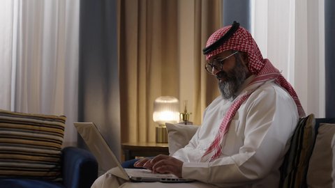 إنجاز مهام العمل عن بعد، مفهوم التقنيات والتكنولوجيا الحديثة المتطورة، رجل عربي خليجي سعودي يرتدي الثوب الابيض و الشماغ يجلس في غرفة المعيشة يقوم باستخدام جهاز الحاسوب المحمول، إجراء مكالمات فيديو عبر الانترنت، ايماءات السعادة و السرور