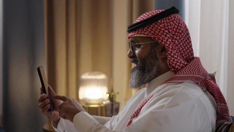 العمل عن بعد وانجاز المهام المطلوبة، استخدام الأجهزة والتقنيات الحديثة، الدردشة والتواصل مع الاصدقاء ، تصوير مقرب من الجانب لرجل عربي خليجي سعودي يرتدي الثوب الابيض و الشماغ يمسك بيده الهاتف المحمول يقوم بتصفح الانترنت ومواقع التواصل الاجتماعي