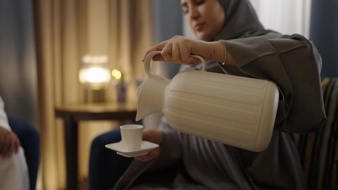 مظهرالنعومه والانوثه، دله نحاسيه وكوب ابيض، ايماءات السعادة و السرور، حسن الاستقبال والكرم عند العرب، إمرأه عربية خليجية سعودية ترتدي الحجاب مع عباءة تجلس علي الاريكة تقوم بقوم بصب الشاي لزوجها