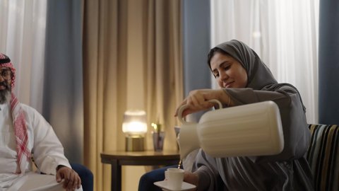 Gestures of happiness and joy, good hospitality and generosity among Arabs, a Gulf Arab Saudi woman wearing a hijab with an abaya sitting on the sofa pouring tea for her husband, a brass coffee pot and a white cup, an appearance of softness and femininity.