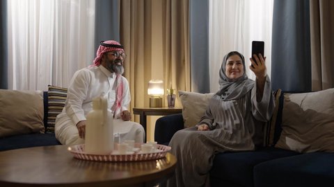 The concept of successful and happy marital relationships, taking commemorative selfies via mobile phone, a Saudi Arabian couple dressed in traditional attire sitting in the living room documenting beautiful moments.