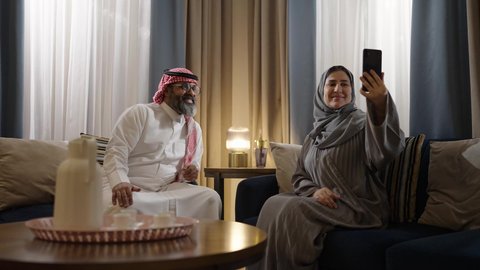 التقاط صور سيلفي تذكارية عبر الهاتف المحمول، مفهوم العلاقات الزوجية الناجحة السعيده، زوجان عربيان خليجيان سعوديان يرتديان الزي التقليدي يجلسان فى غرفة المعيشة يقومان بتوثيق اللحظات الجميلة
