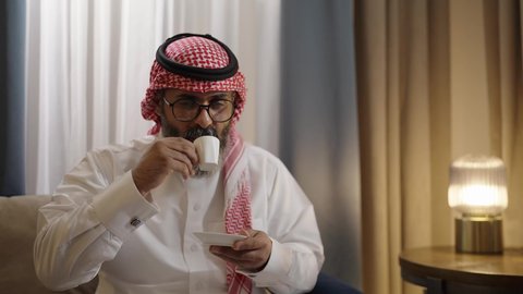 قضاء أوقات ممتعة ورومانسية، زوجان عربيان خليجيان سعوديان مبتسمان يجلسان في  غرفه المعيشة ، مفهوم المودة والالفة ، ايماءات السرور والسعادة ،الاستمتاع بشرب المشروبات الساخنه