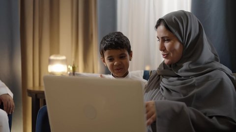 تصفح مواقع التواصل الاجتماعي عبر الهاتف المحمول، قضاء أوقات ممتعة، أنشطة ترفيهية الكترونية مسلية، التعلم عن بعد وتلقي الدروس عبر الأونلاين، استخدام التكنولوجيا والتقنية الحديثة والمتطورة، إمرأة عربية خليجية اماراتية ترتدي الحجاب والعباءة تجلس برفقة ابنها و زوجها وتستخدم جهاز الحاسوب المحمول