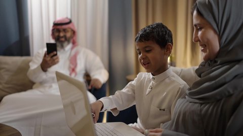 أنشطة ترفيهية الكترونية مسلية، التعلم عن بعد وتلقي الدروس عبر الأونلاين، استخدام التكنولوجيا والتقنية الحديثة والمتطورة، قضاء أوقات ممتعة، إمرأة عربية خليجية اماراتية ترتدي الحجاب والعباءة تجلس برفقة ابنها و زوجها وتستخدم جهاز الحاسوب المحمول، تصفح مواقع التواصل الاجتماعي عبر الهاتف المحمول