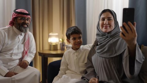 Building a growing Saudi community, family gathering in the living room, the joy of children spending quality time with their parents, taking a memorable family selfie via mobile phone, a smiling Arab Saudi Gulf family documenting enjoyable moments with gestures of happiness and joy.