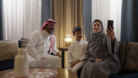 التقاط سيلفي عائلي تذكاري عبر الهاتف المحمول, فرحة الأبناء بقضاء وقت ممتع مع الأهل, عائلة عربية سعودية خليجية مبتسمة تقوم بتوثيق اللحظات الممتعة بإيماءات السعادة والفرح, الاجتماع الأسري في غرفة المعيشة وبناء مجتمع سعودي متنامي
