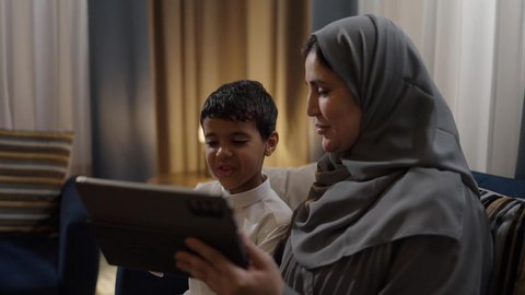 قضاء أوقات مليئه بالحب والموده مع الابناء، امراه عربيه خليجيه سعودية ترتدي العباءة و الحجاب تجلس برفقة ابنها تقوم بمشاهدة المسلسلات الممتعه عبر جهاز التابلت، متابعه مواقع التواصل الاجتماعي، استخدام الأجهزة الالكترونيه والتقنيات الحديثه، أجواء عائليه منزليه هادئه