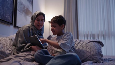 المشاركه بقراءه القصص المفيده مع الأبناء، مفهوم التربيه بالحب والحنان، امرأة عربية خليجية سعودية ترتدي العباءه والحجاب تقرأ الروايات المسليه لابنها في غرفه النوم عبر جهاز التابلت, استغلال الاوقات في تعليم وتربيه الاولاد، استخدام جهاز تقني حديث