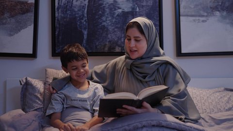 قراءة القصص والروايات، ام تقضي وقت ممتع مع الابن قبل النوم، اهتمام الأم بالابناء، امرأة عربية خليجية سعودية ترتدي العباءة و الحجاب تقرأ لإبنها قصه مسلية، مفهوم السعادة الاسرية،لأنشطة التعليمية وتنميه قدرات الابناء بالتعلم