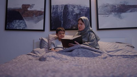 ام تقضي وقت ممتع مع الابن قبل النوم، اهتمام الأم بالابناء، امرأة عربية خليجية سعودية ترتدي العباءة و الحجاب تقرأ لإبنها قصه مسلية، قراءة القصص والروايات، مفهوم السعادة الاسرية،لأنشطة التعليمية وتنميه قدرات الابناء بالتعلم