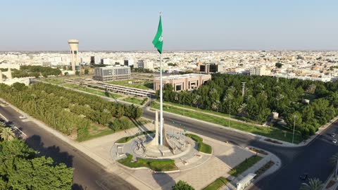 Pride and honor in belonging to the homeland, a drone shot of the green Saudi flag waving in the air in the city of Tabuk, the famous landmarks in the Kingdom of Saudi Arabia, and the traffic of cars in the streets of the city.