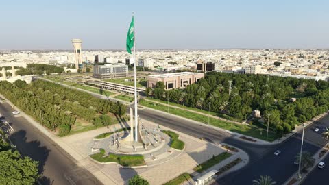 Pride and honor in belonging to the homeland, a drone shot of the green Saudi flag waving in the air in the city of Tabuk, the famous landmarks in the Kingdom of Saudi Arabia, and the traffic of cars in the streets of the city.