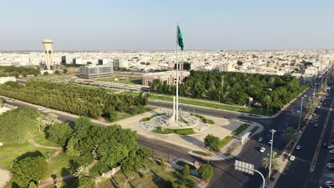 الفخر والاعتزاز بالانتماء الى الوطن,تصوير درون لسارية العلم السعودي الأخضر المرفرف في الهواء في مدينة تبوك،  المعالم المشهورة في المملكة العربية السعودية ،حركة مرور السيارات في شوارع المدينة