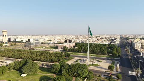 Pride and honor in belonging to the homeland, a drone shot of the green Saudi flag waving in the air in the city of Tabuk, the famous landmarks in the Kingdom of Saudi Arabia, and the traffic of cars in the streets of the city.
