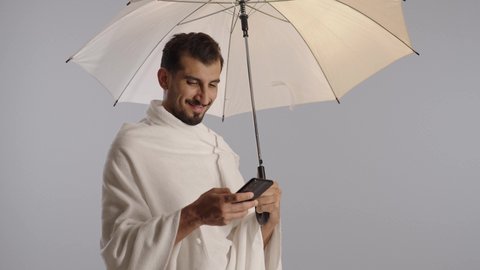 The concept of the pillars of Islam, the electronic Hajj guide application, the use of the guidance application service to perform the rituals of Hajj and Umrah, a portrait of a Gulf Arab Muslim man from Saudi Arabia wearing Ihram clothing and holding a white umbrella while using a mobile phone, gray background.