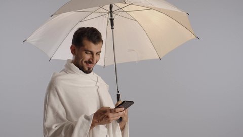 تطبيق مرشد الحج الالكتروني، استخدام خدمة تطبيق الارشادات لأداء مناسك الحج والعمرة، بورتريه لرجل مسلم خليجي عربي سعودي يرتدي ملابس الإحرام و يمسك شمسية بيضاء يستخدم الهاتف المحمول، مفهوم أركان الإسلام، خلفية رمادية